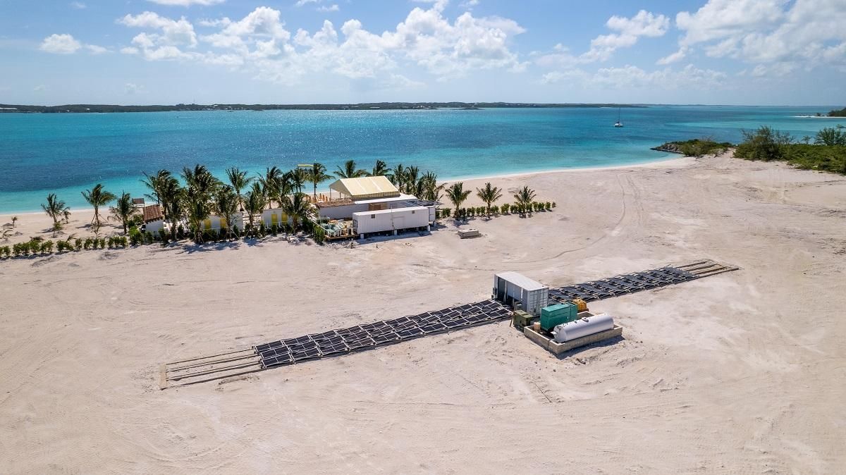 Aerial view of a sandy beach with a building and long lines of dark barriers in front of blue water.
