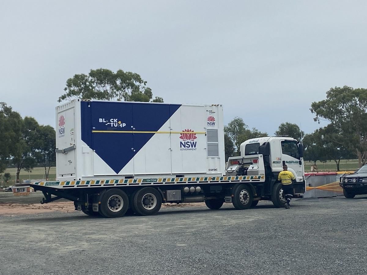A white truck with a blue and white box on its flatbed, parked on gravel. A person stands nearby.