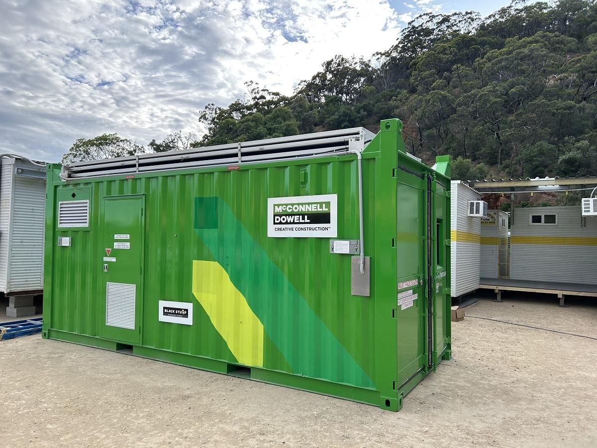 Green container with branding, outdoors. Cloudy sky and hill in the background.