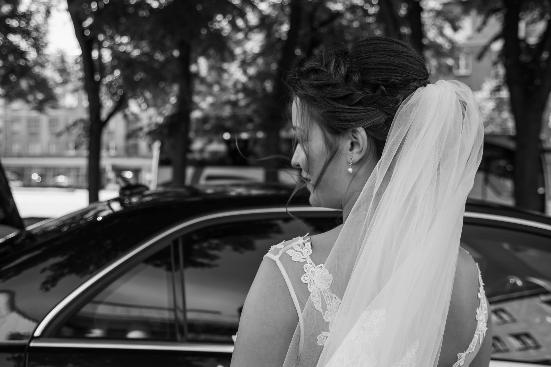 Couple in wedding attire exiting a white car; bride holding bouquet, groom's arm around her.