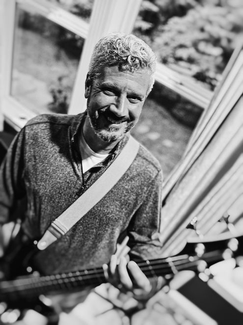 Man with gray hair and beard smiles, holding a bass guitar. He wears a sweater and stands near a window.