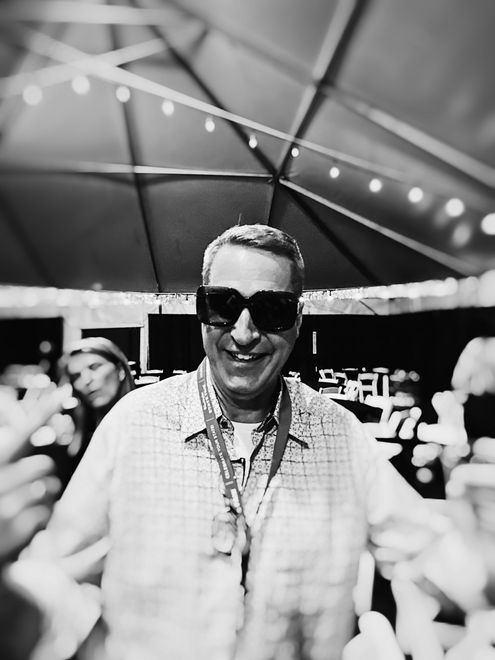 Man wearing sunglasses smiles at the camera, under a tent with string lights; people in background.
