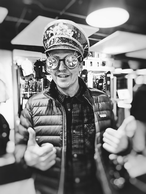 Man wearing captain's hat and novelty sunglasses, giving thumbs up, smiling. In a shop.