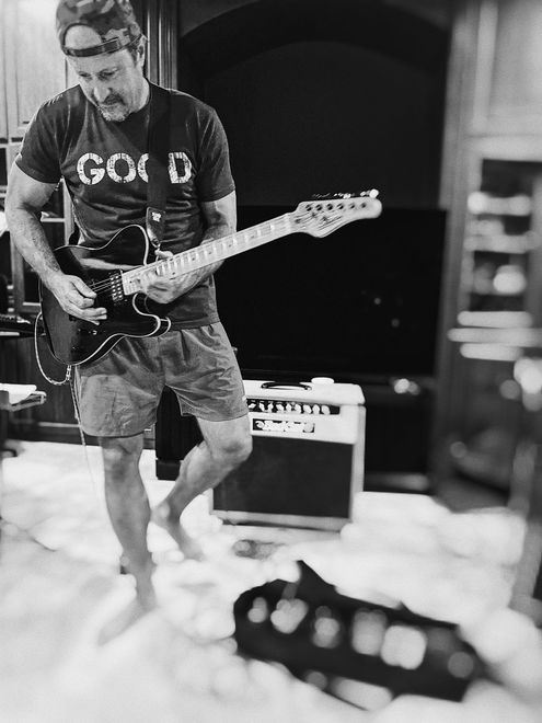 Man playing guitar barefoot indoors. Wearing a “GOOD” shirt and shorts, amplifier behind him.