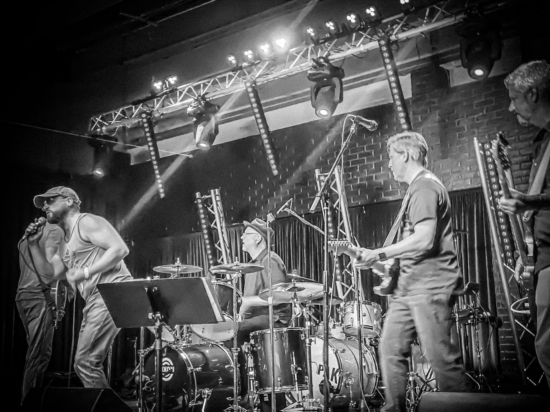 Band performing on stage, with singer, drummer, guitarist, and other musicians under stage lights.