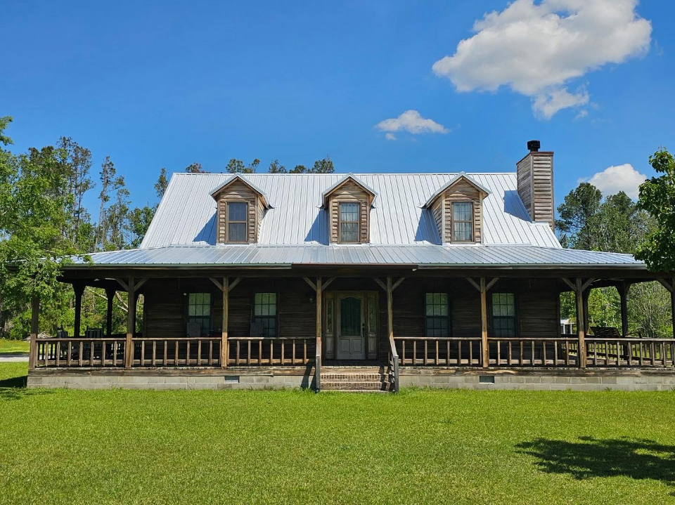 Historic white farmhouse with porch and dormer windows under a blue sky, surrounded by green lawn.