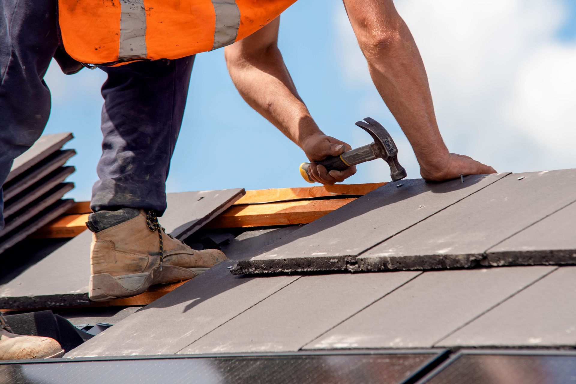 Roofing worker nailing gray metal roof panels with a hammer, wearing orange safety vest and boots.