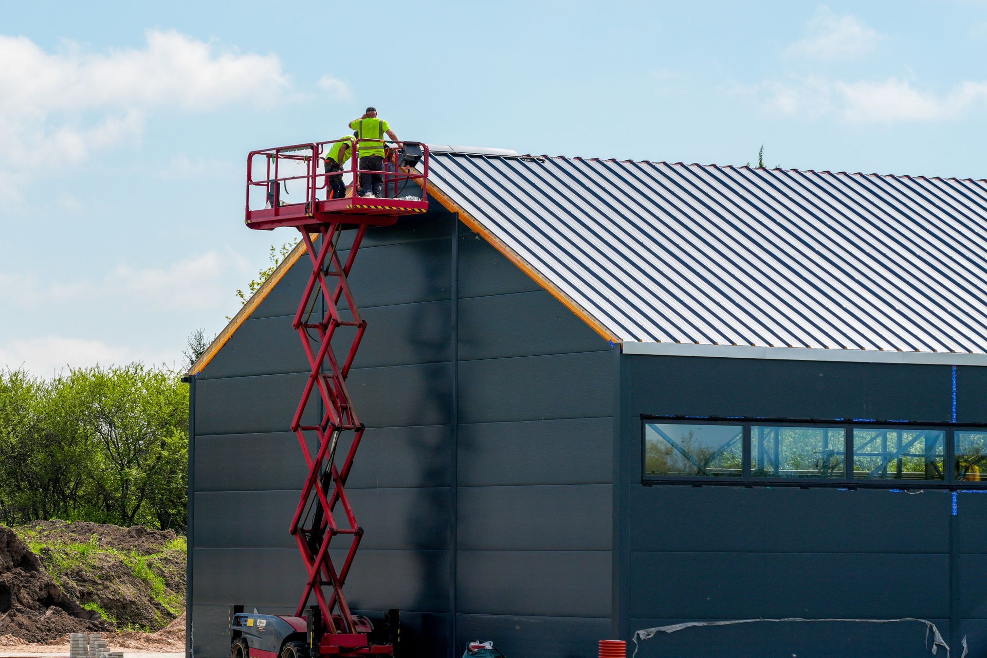 Workers on a red scissor lift repairing the roof of a gray industrial building
