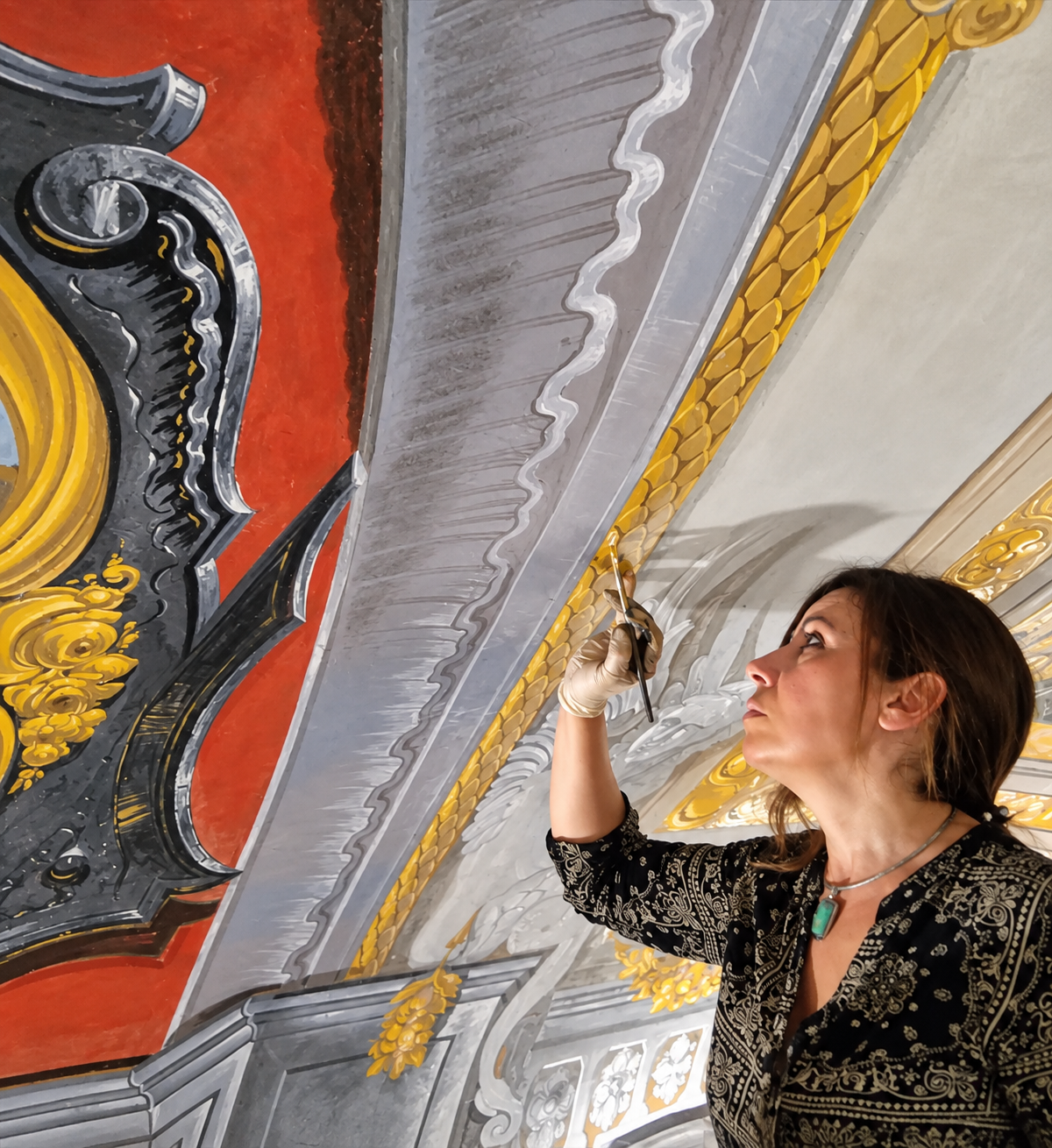 Donna in abito nero dipinge con un pennello i dettagli decorati del soffitto, aggiungendo accenti dorati.