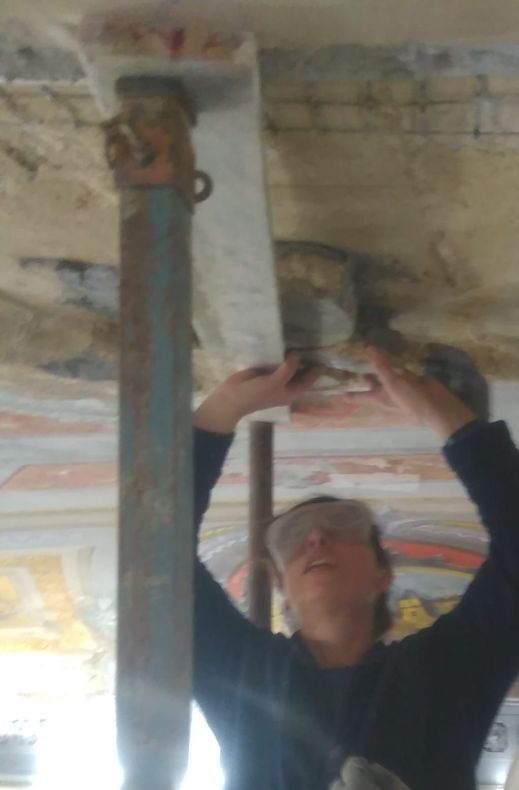 Una persona al lavoro sopra la testa su una trave del soffitto, con un attrezzo in mano, in una stanza con modanature dipinte