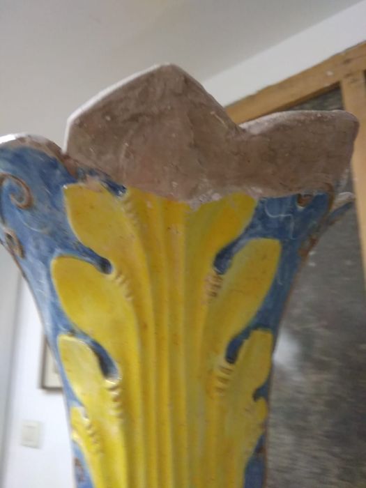 Colonna ornamentale colorata con scanalature gialle e dettagli blu, sormontata da un capitello grezzo marrone, in un ambiente interno