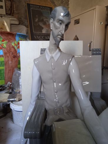 Scultura umanoide grigia seduta al chiuso, con una camicia bianca e il busto grigio lucido, in una stanza disordinata