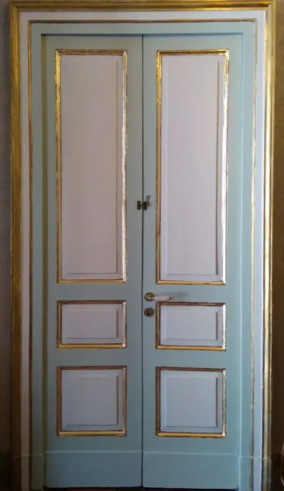 Doppia porta azzurro pastello con finiture e pannelli dorati, chiusa all’interno di un vano porta incorniciato