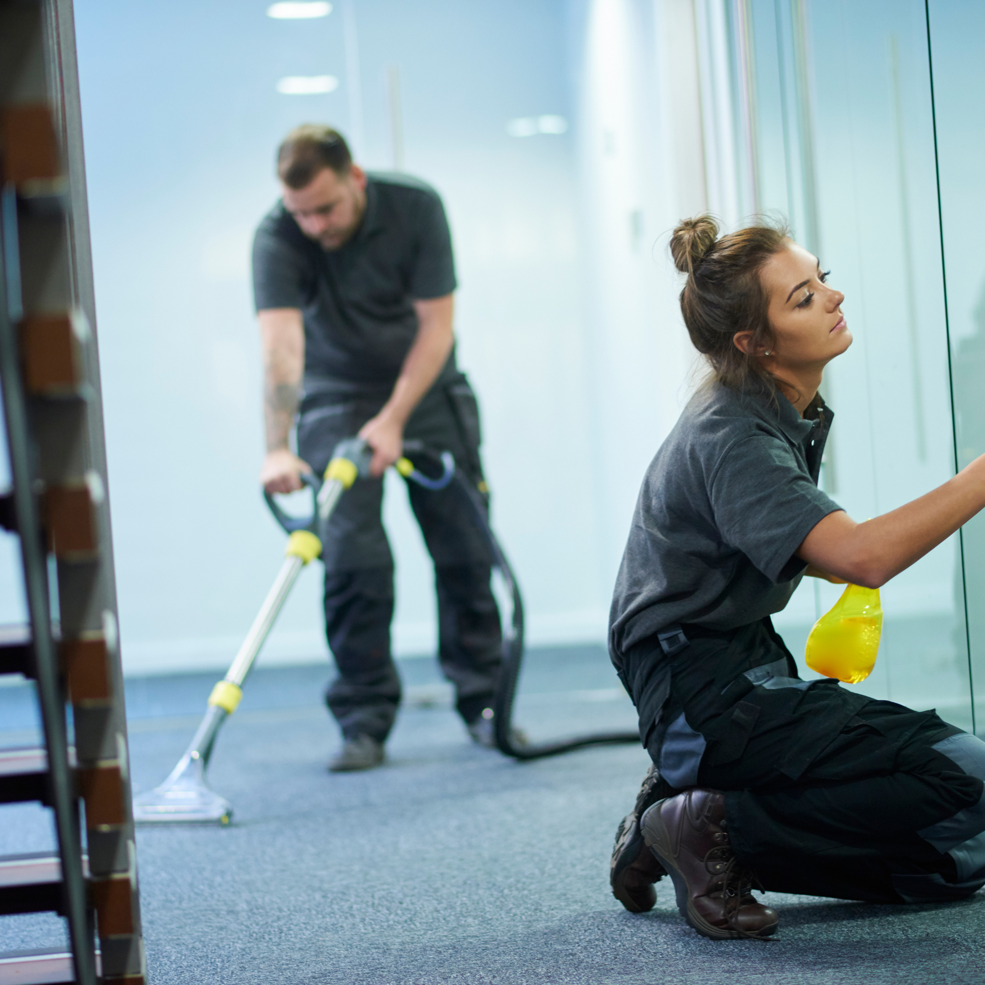 Two cleaners in an office: one vacuums carpet, the other cleans glass.