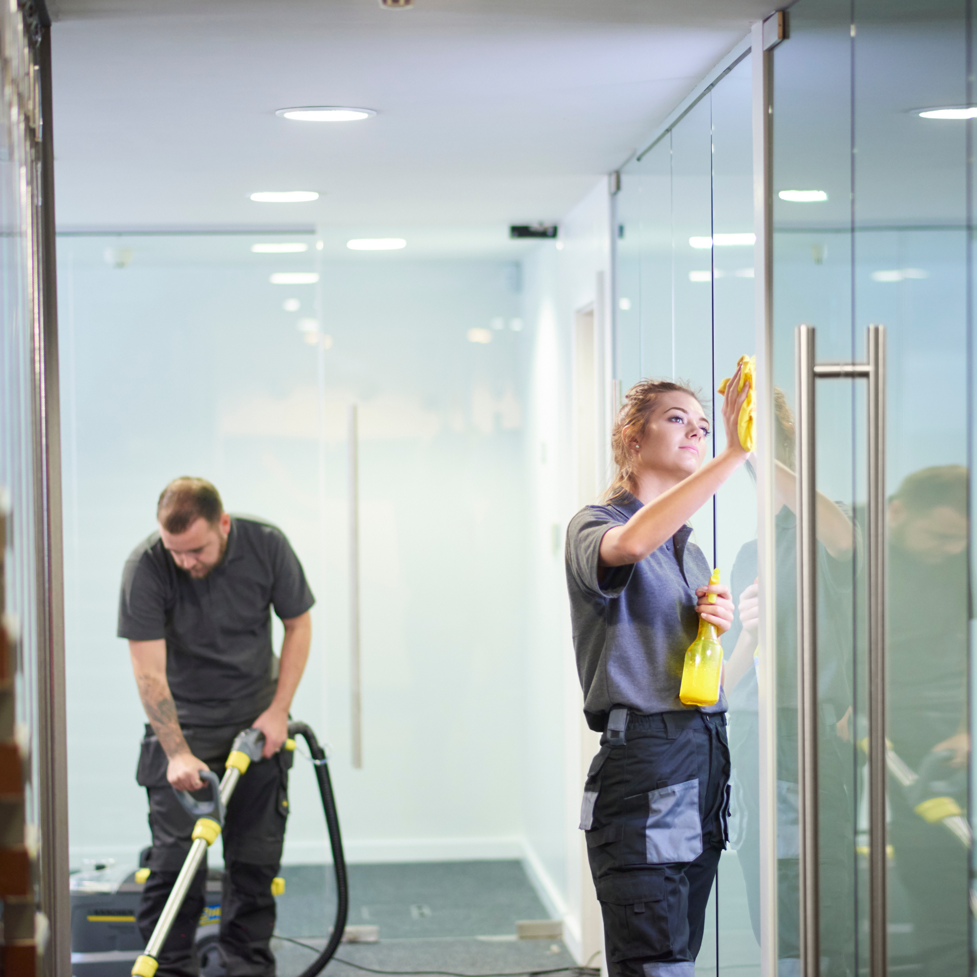 Two people cleaning an office. One vacuums, the other wipes glass doors with a yellow cloth and spray bottle.