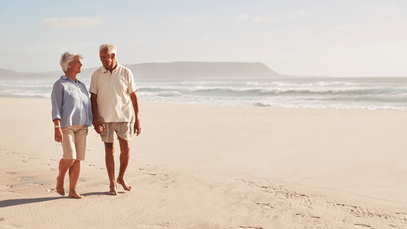 An elderly couple is walking on the beach holding hands.