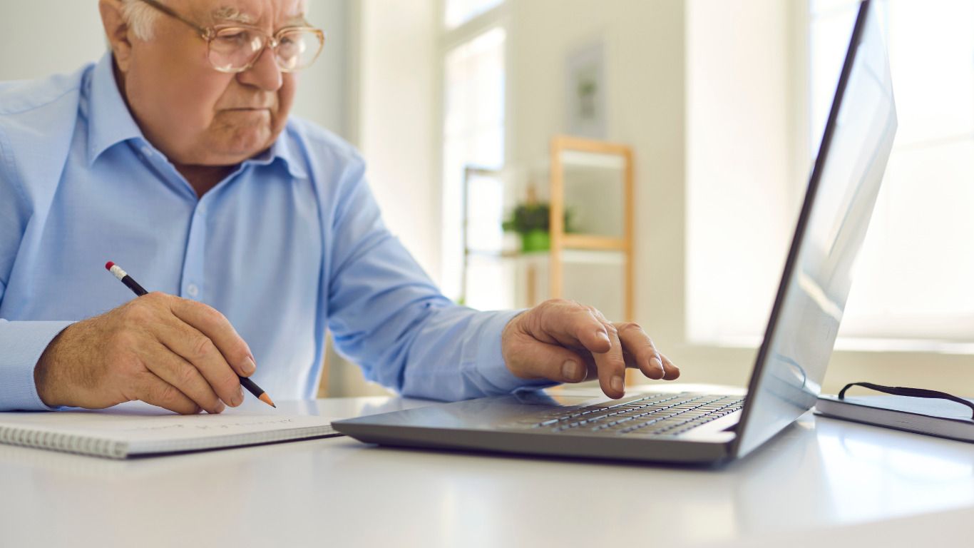 An elderly man is sitting at a desk using a laptop computer.