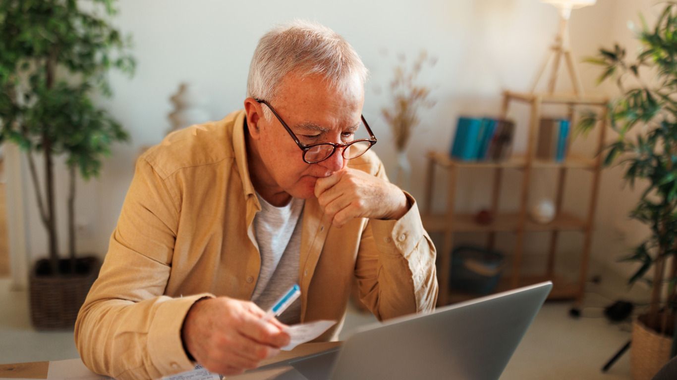 An elderly man is sitting at a table using a laptop computer.