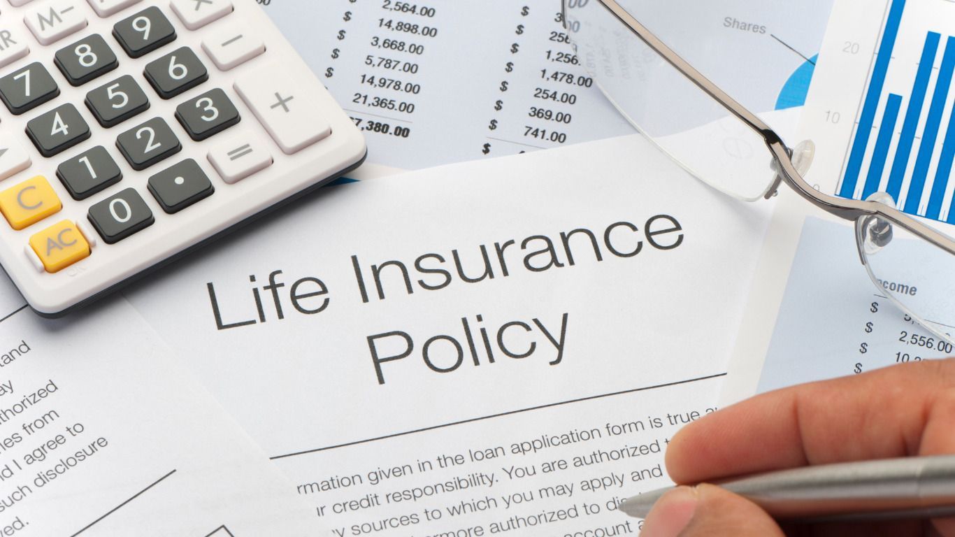 A person is writing on a life insurance policy next to a calculator and glasses.