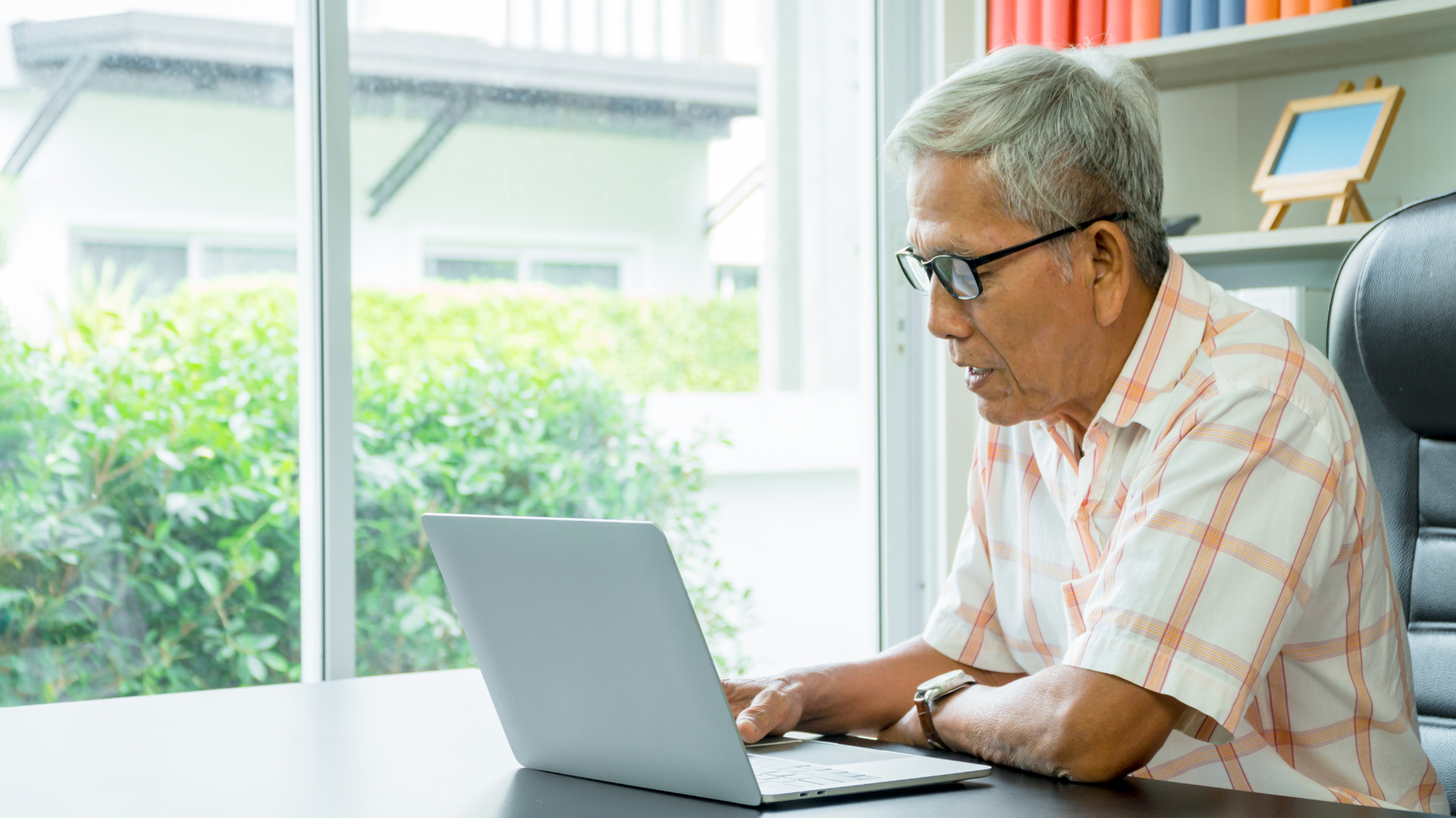 An elderly man is sitting at a desk using a laptop computer.