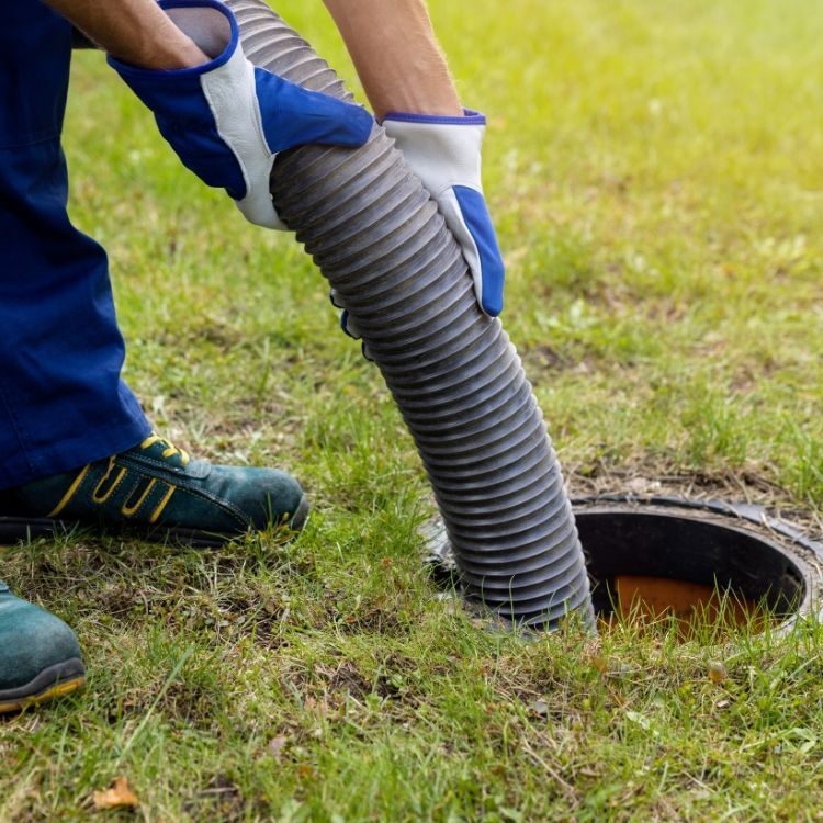 Gloved worker lowering a corrugated hose into a round ground opening on a grassy lawn