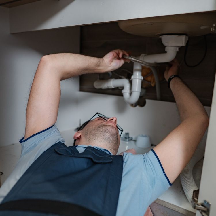 Plumber repairing pipes under a sink cabinet, reaching with tools in a tight space.