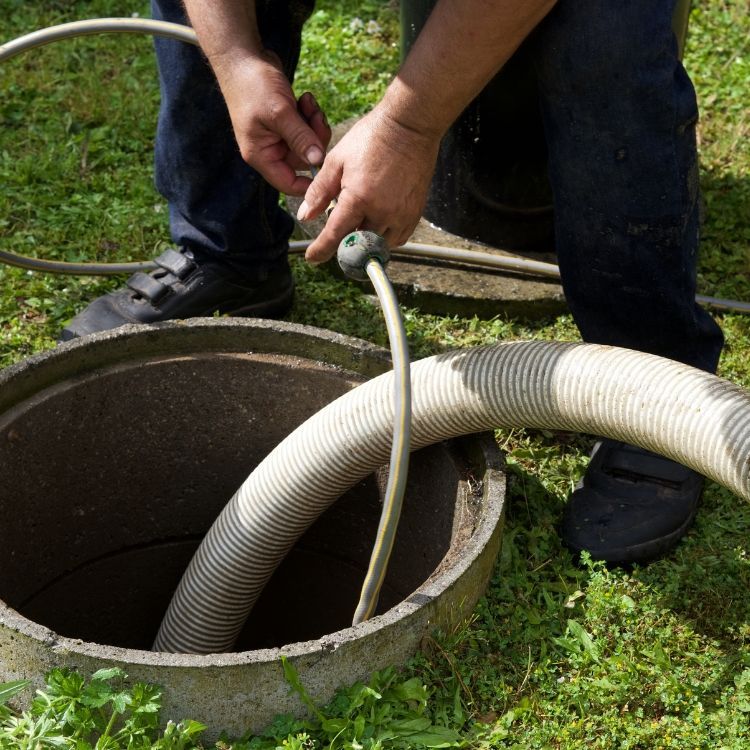Person lowering a white hose into a round concrete drain opening in a grassy yard