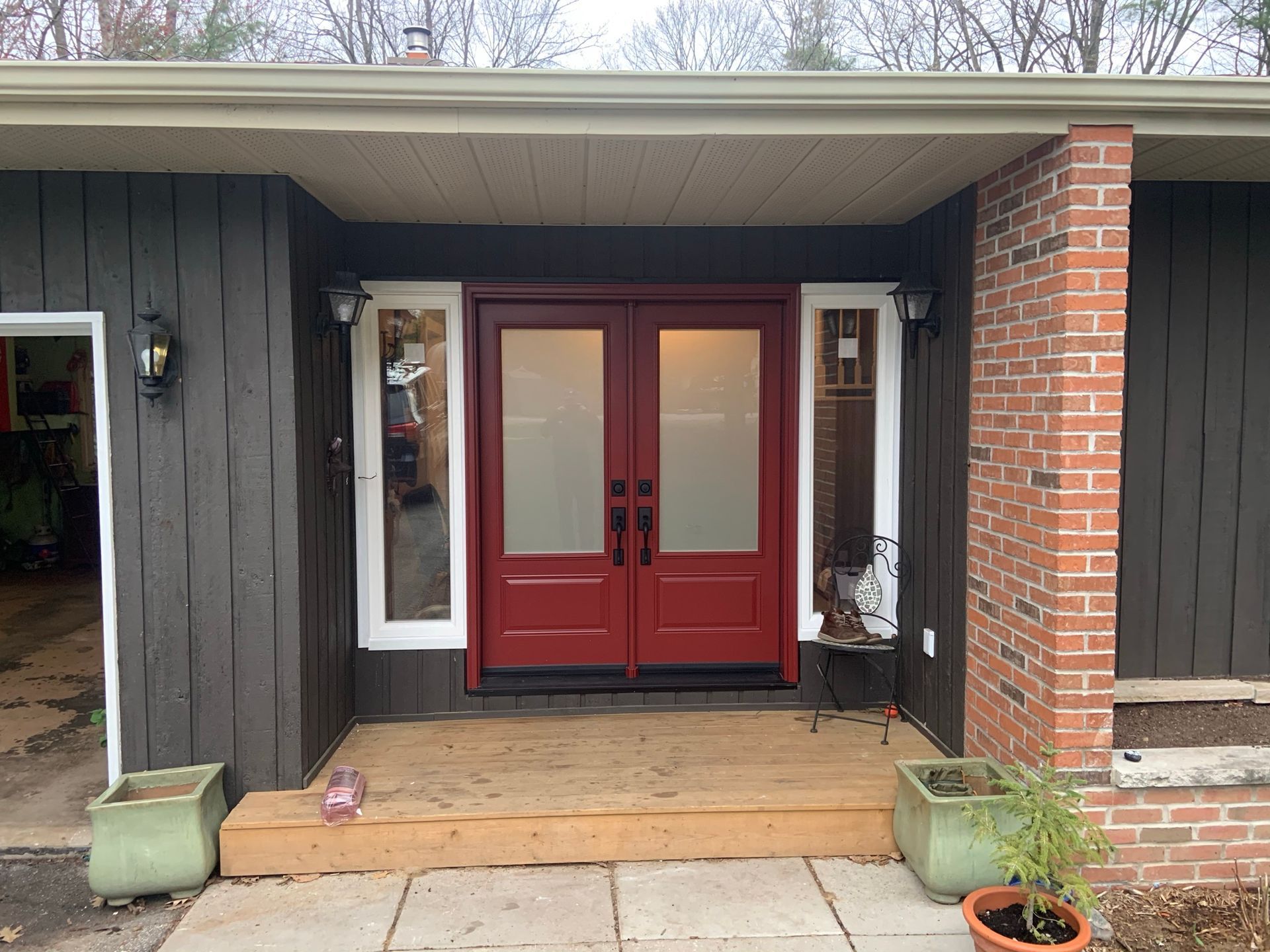 Front porch with red double doors, brick columns, dark siding, and potted plants on either side.