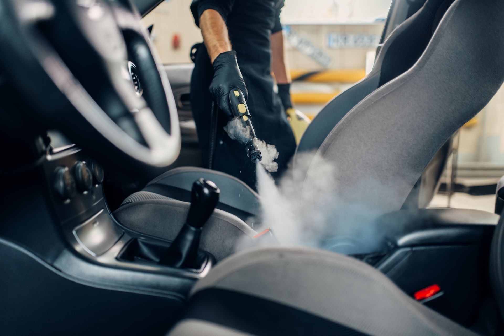 Persona che pulisce a vapore l'interno di un'auto, concentrandosi sui sedili, generando vapore visibile.
