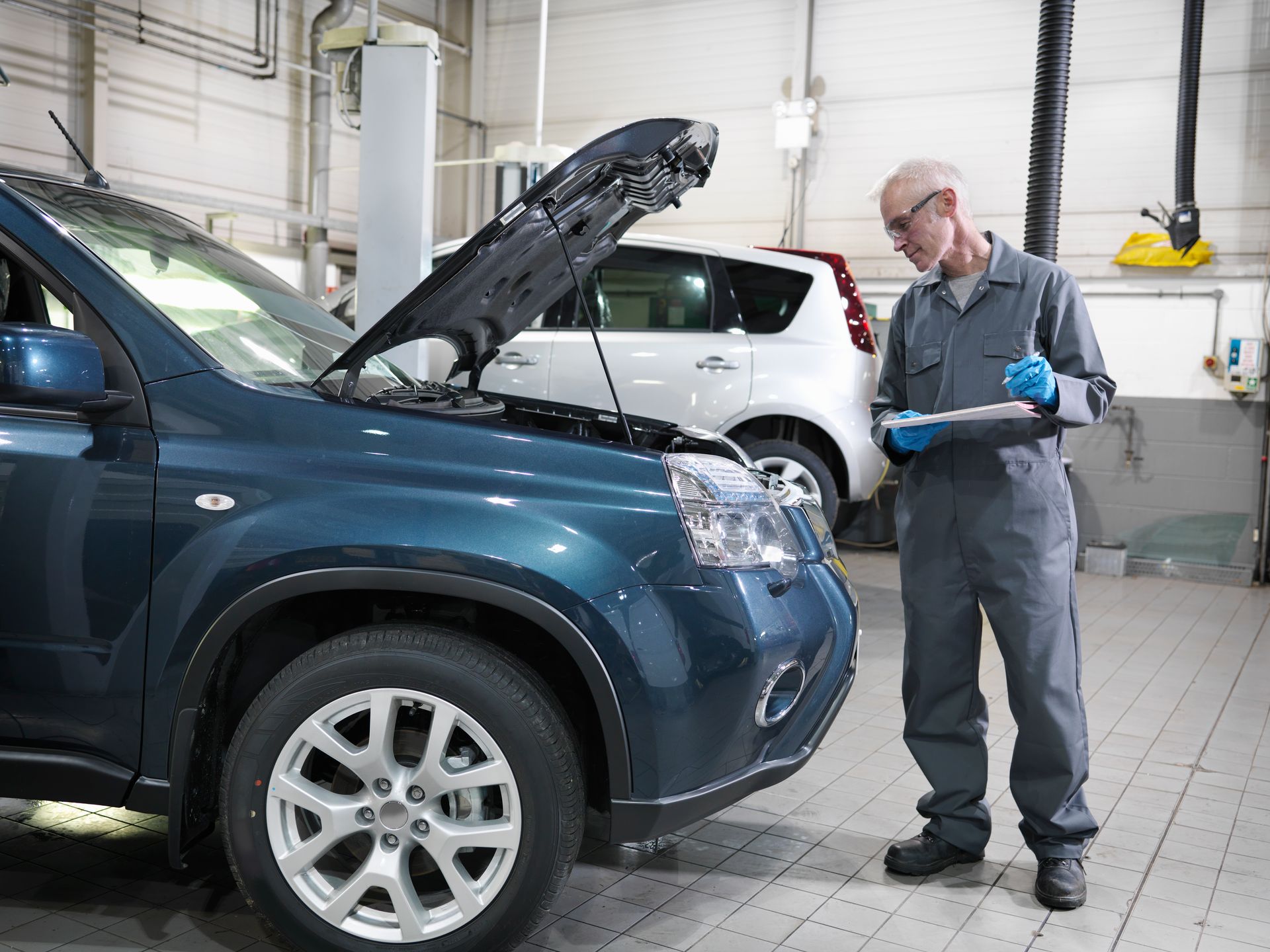 Meccanico in tuta blu che ispeziona un SUV blu con il cofano aperto in un'officina, mentre guarda un blocco per appunti.