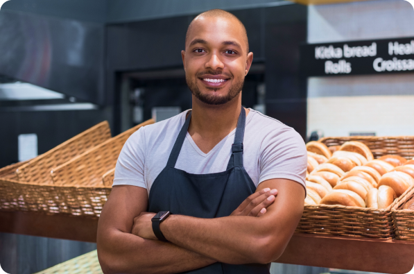 Padeiro sorridente com os braços cruzados em uma padaria ao lado de cestas de pão e croissants.