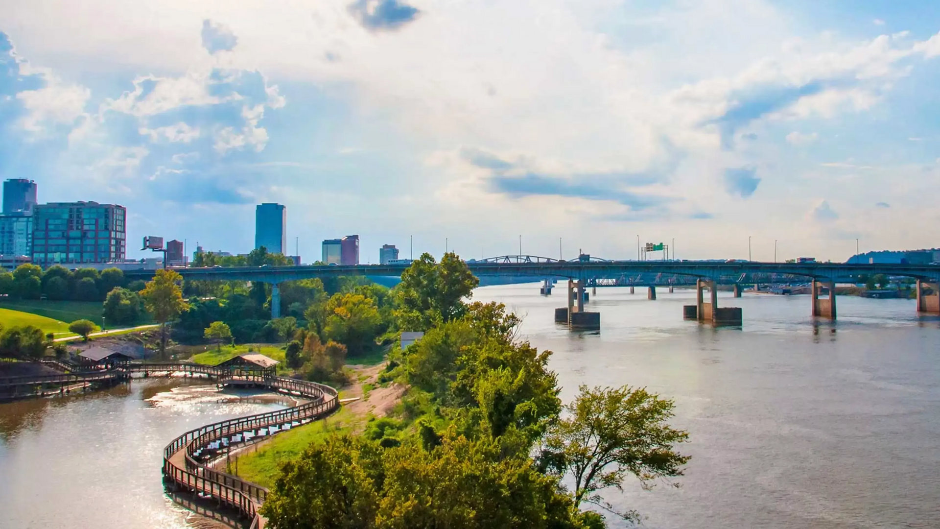 A wide shot of Little Rock, Arkansas, cityscape with river and bridge under a cloudy sky.