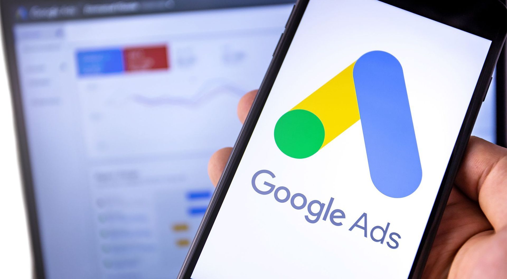 Hand holding a phone displaying the Google Ads logo against a blurred background of a computer screen with charts.