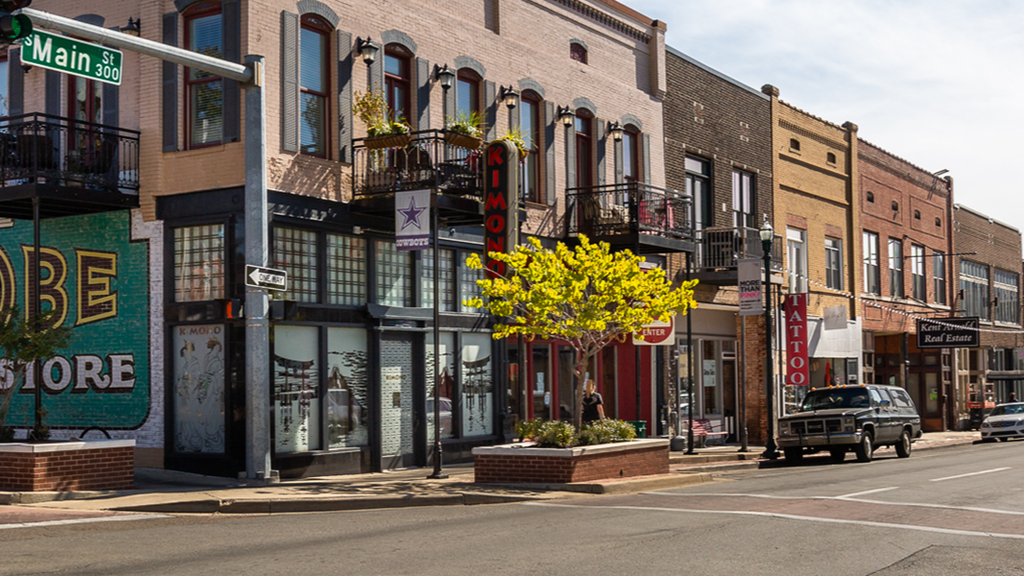 Street with brick buildings, storefronts, parked vehicles, and a street sign.