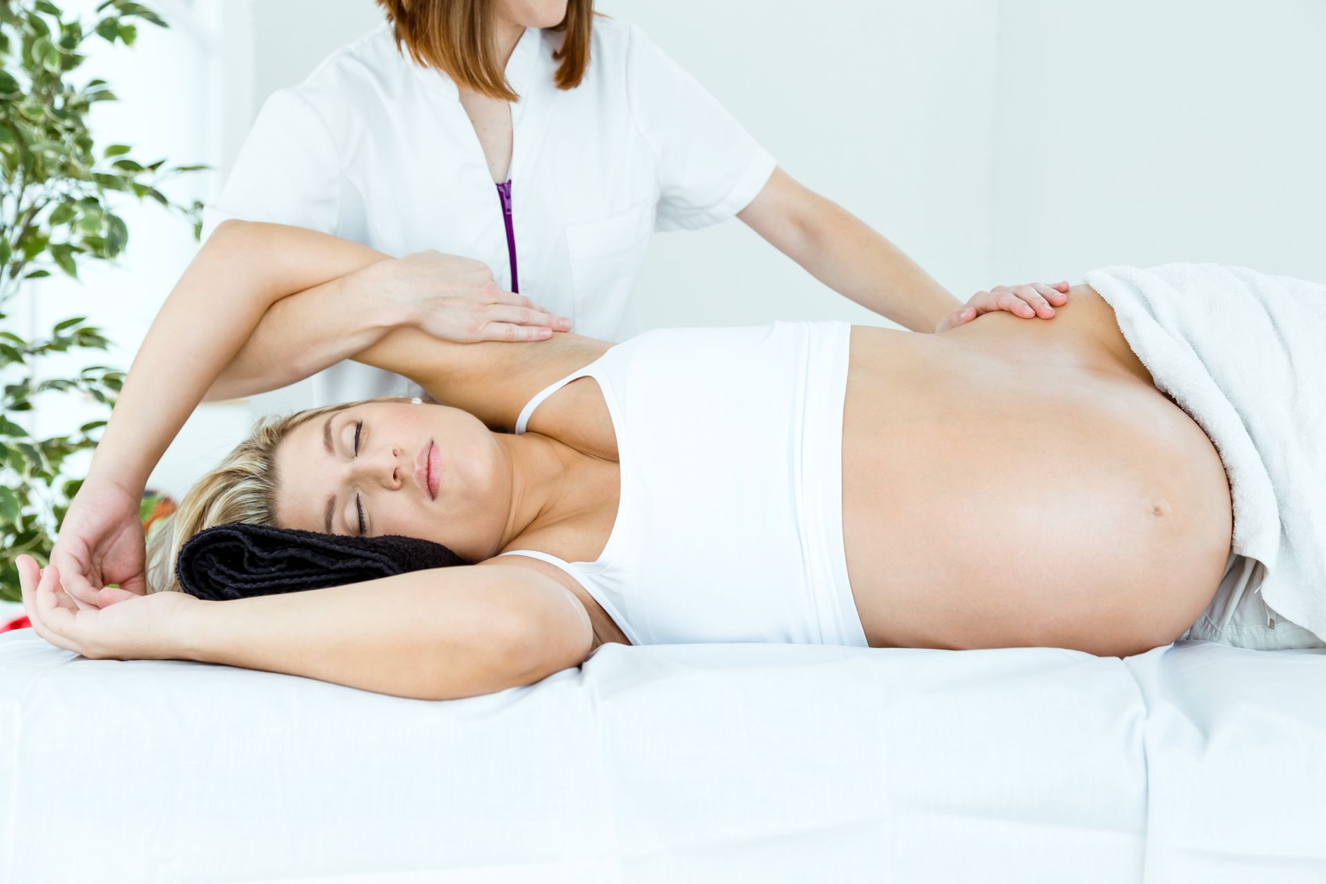 Two women on a white bed, one massaging the other’s back in a bright bedroom Two women on a white bed, one massaging the other’s back in a bright bedroom