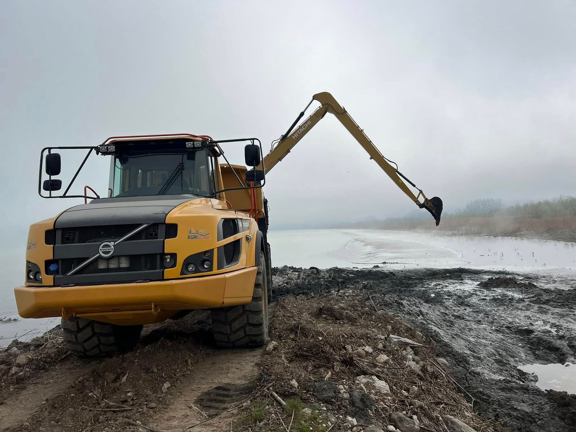 Un camion Volvo giallo con un braccio escavatore al lavoro in riva al mare, in una giornata nuvolosa.