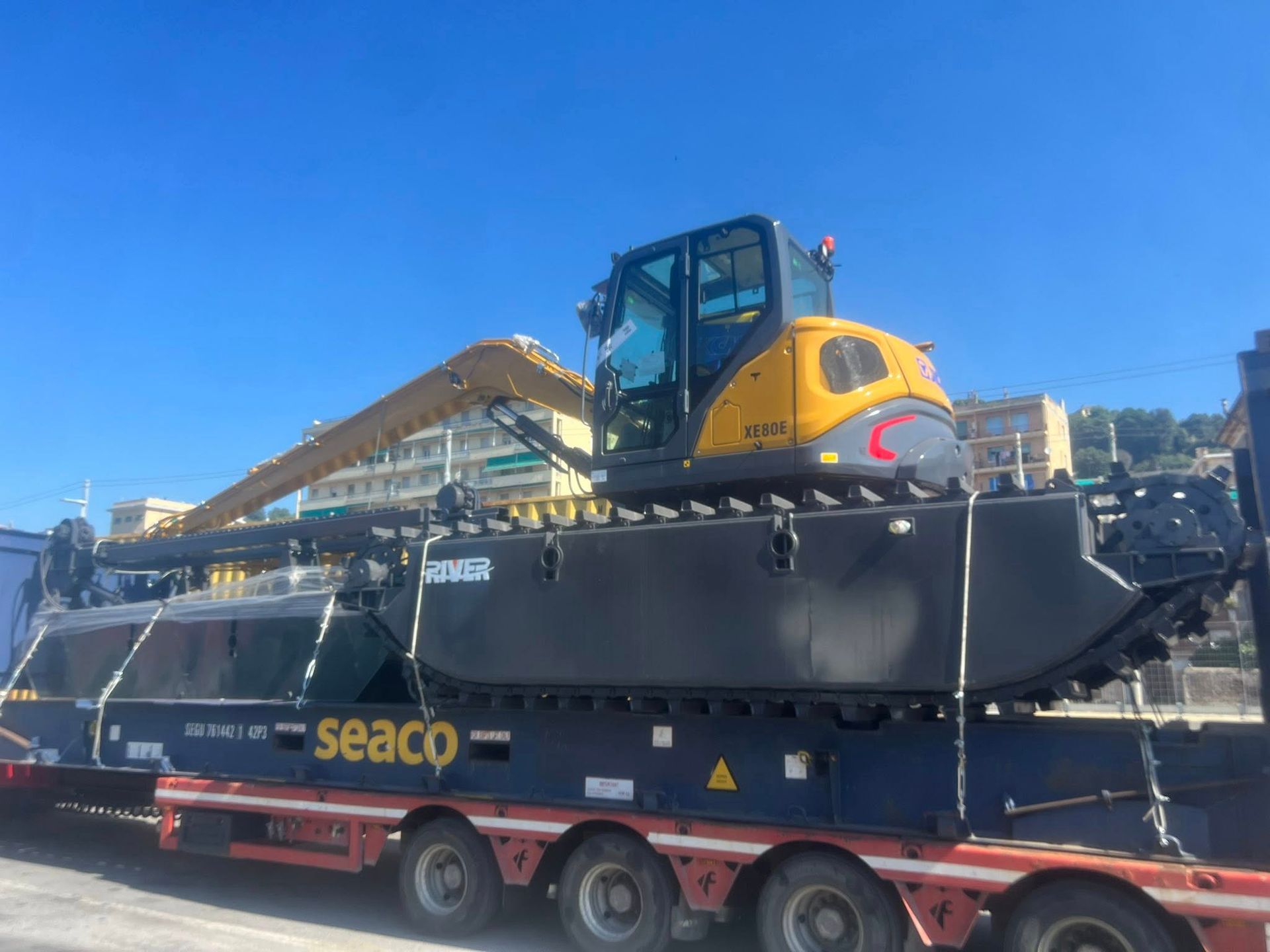 Escavatore giallo su cingoli anfibi neri, caricato su un camion da trasporto, sotto un cielo azzurro.
