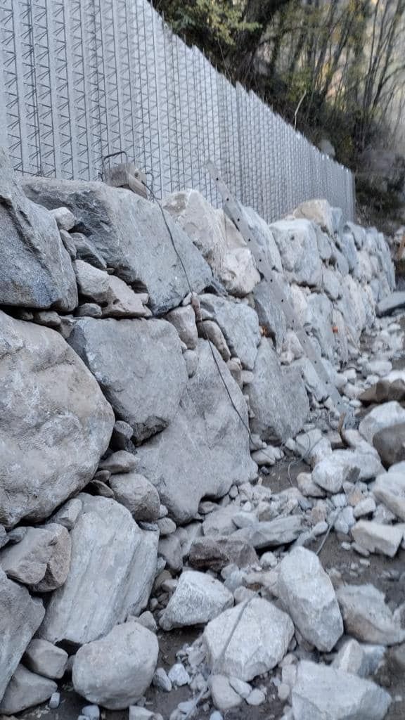 Muro di pietra di fronte a un muro di contenimento in blocchi di cemento. Grandi rocce e detriti sono sparsi in primo piano.