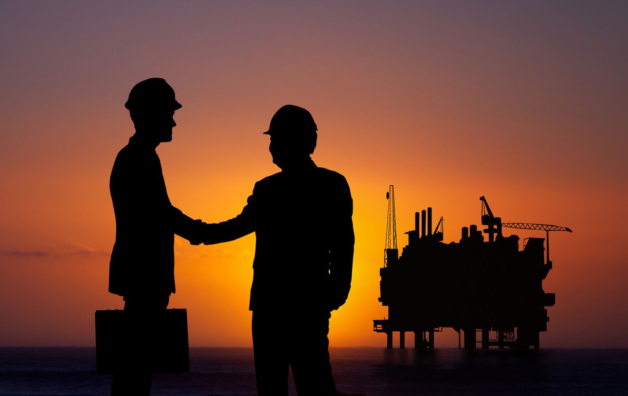 Two silhouetted workers shake hands at sunset beside an offshore oil rig.