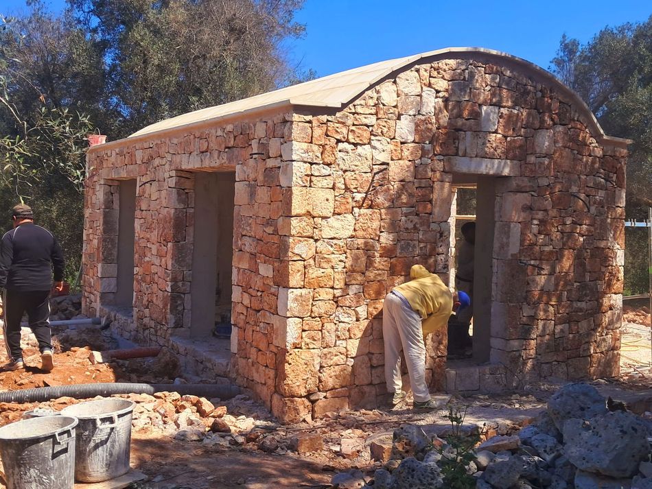 Piccolo edificio in pietra in costruzione, con operai e macerie sparse all’aperto