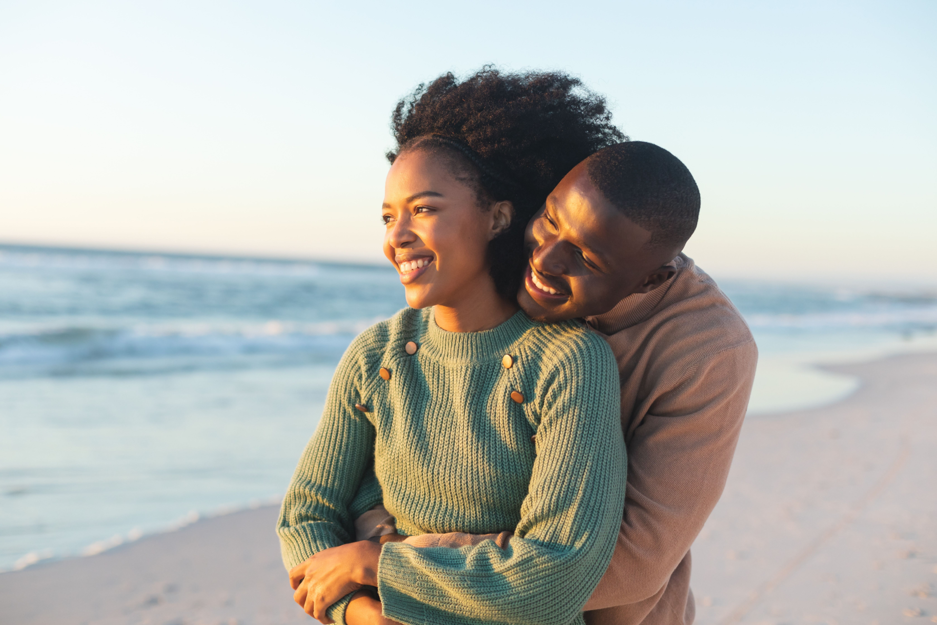Couple hugging on a beach; woman in green sweater smiling, man in tan sweater smiling from behind her; ocean background.