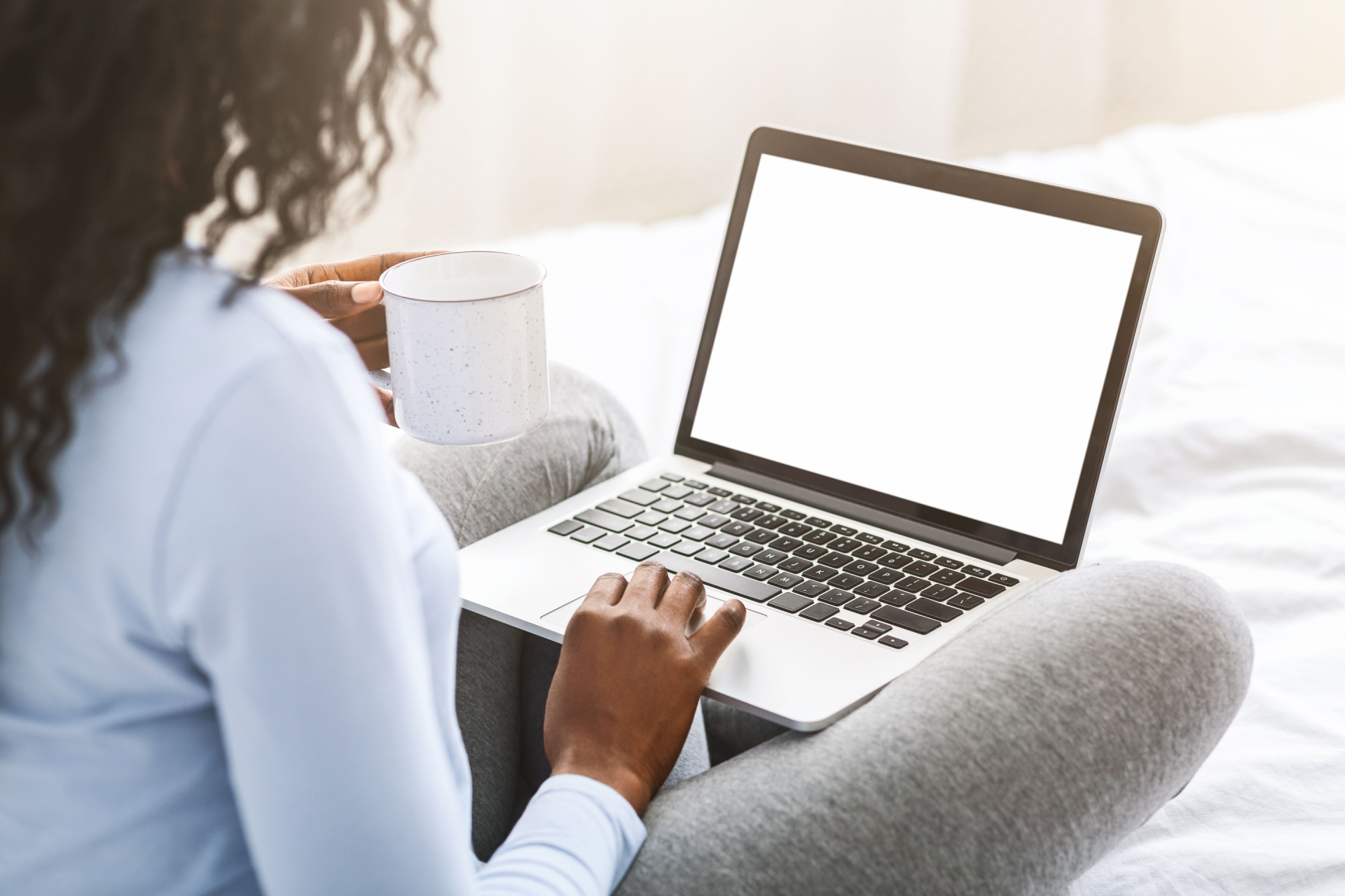 Person using a laptop, holding a mug, sitting on a bed.