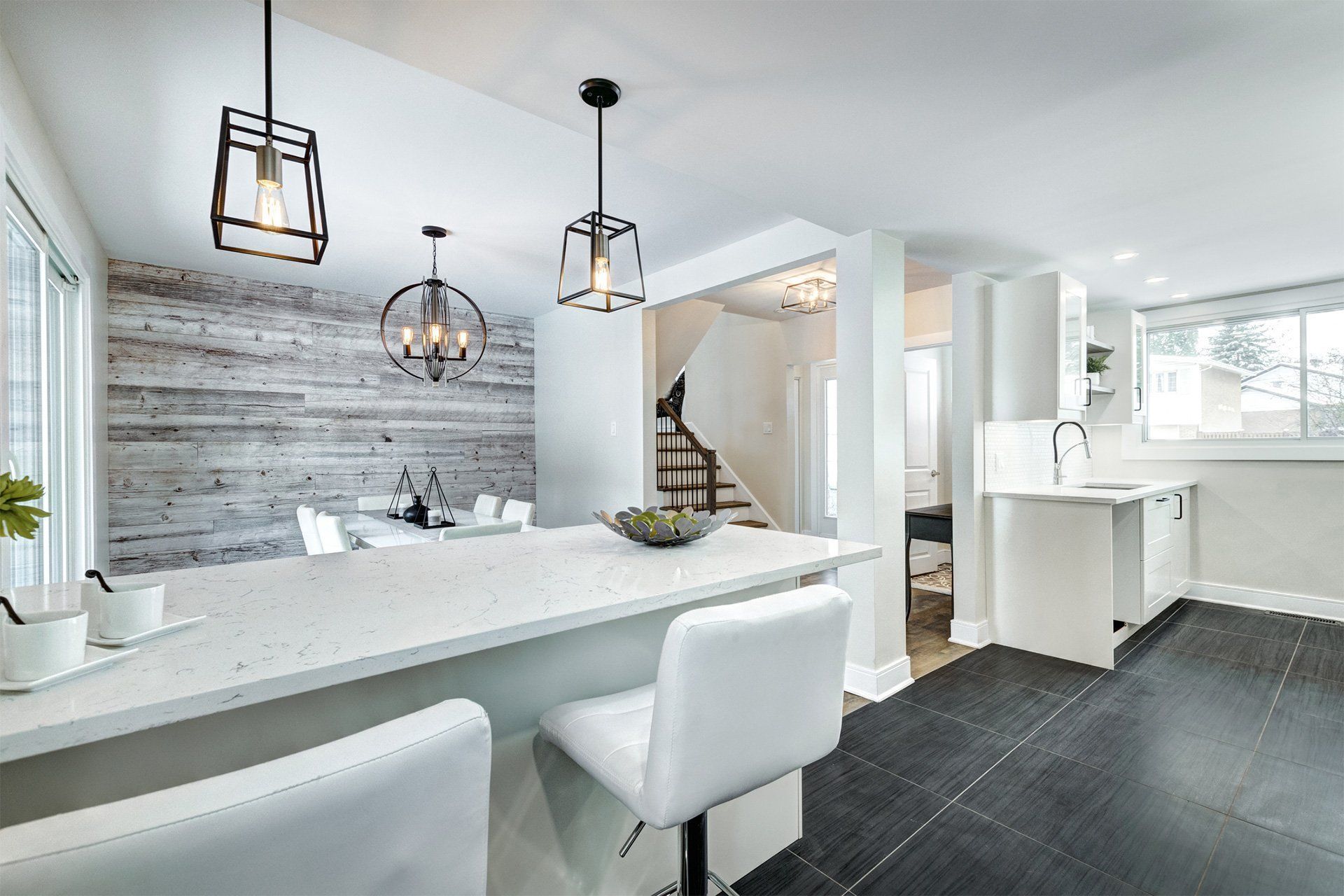 Modern kitchen and dining area with white countertops, gray wood wall, and black light fixtures.
