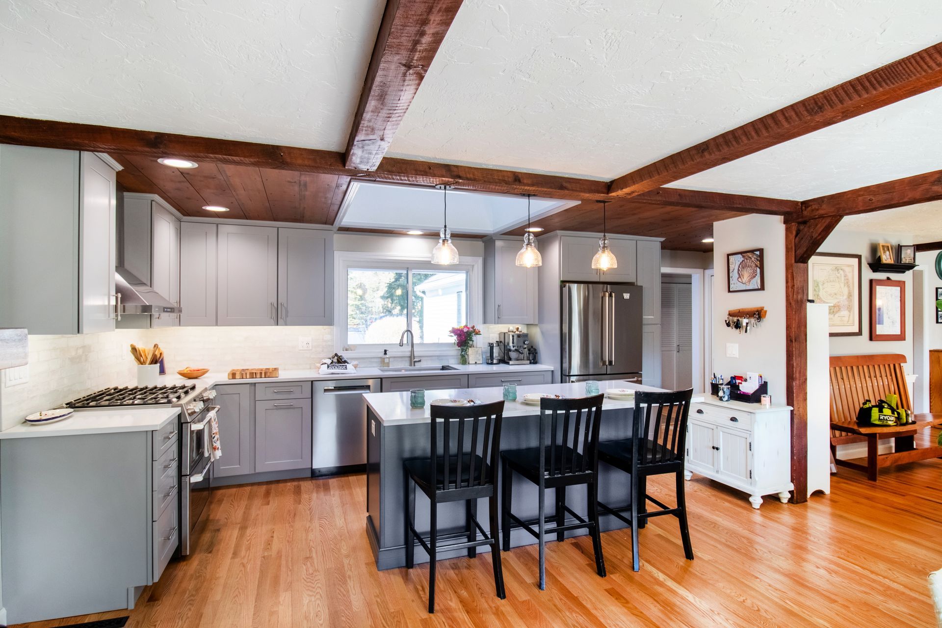 Kitchen with gray cabinets, stainless steel appliances, wood beams, island with black chairs, and hardwood floors.