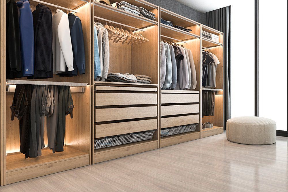 Walk-in closet with lit shelves, hanging clothes, drawers, and a beige ottoman in a bright room.