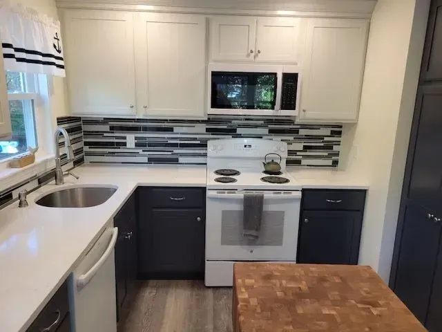 A kitchen with white countertops, dark blue lower cabinets, and a white oven.  Backsplash is a mix of gray and white tiles.