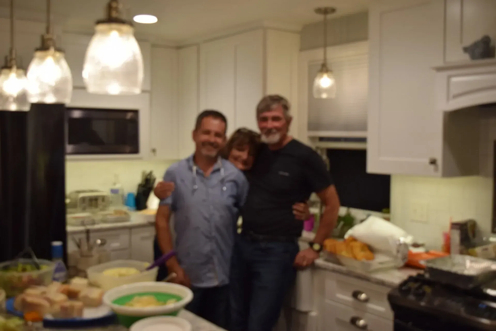Three people smiling in a bright kitchen. One woman is sandwiched between two men, arms around each other.