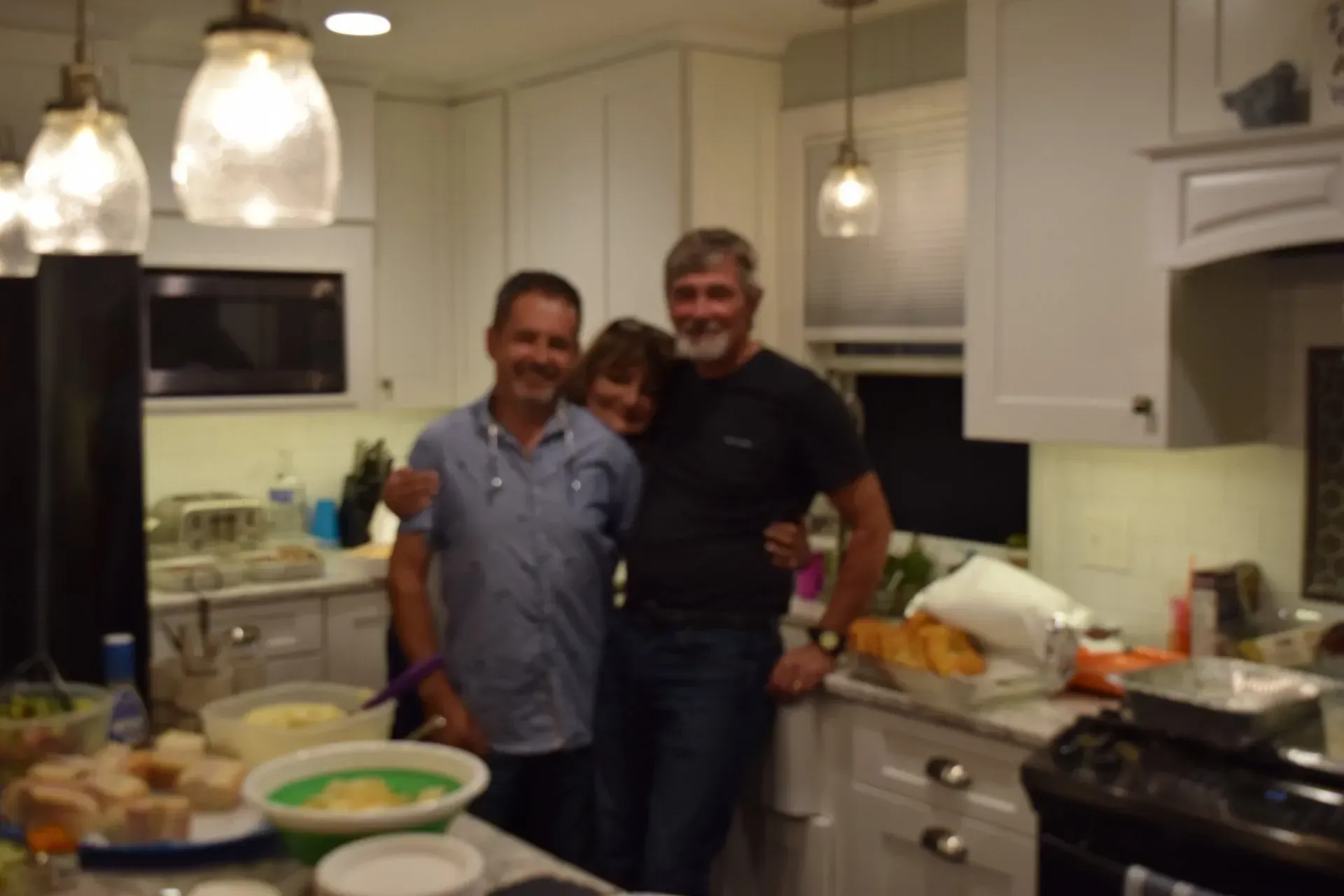 Three people smiling in a bright kitchen. One woman is sandwiched between two men, next to food preparation areas.