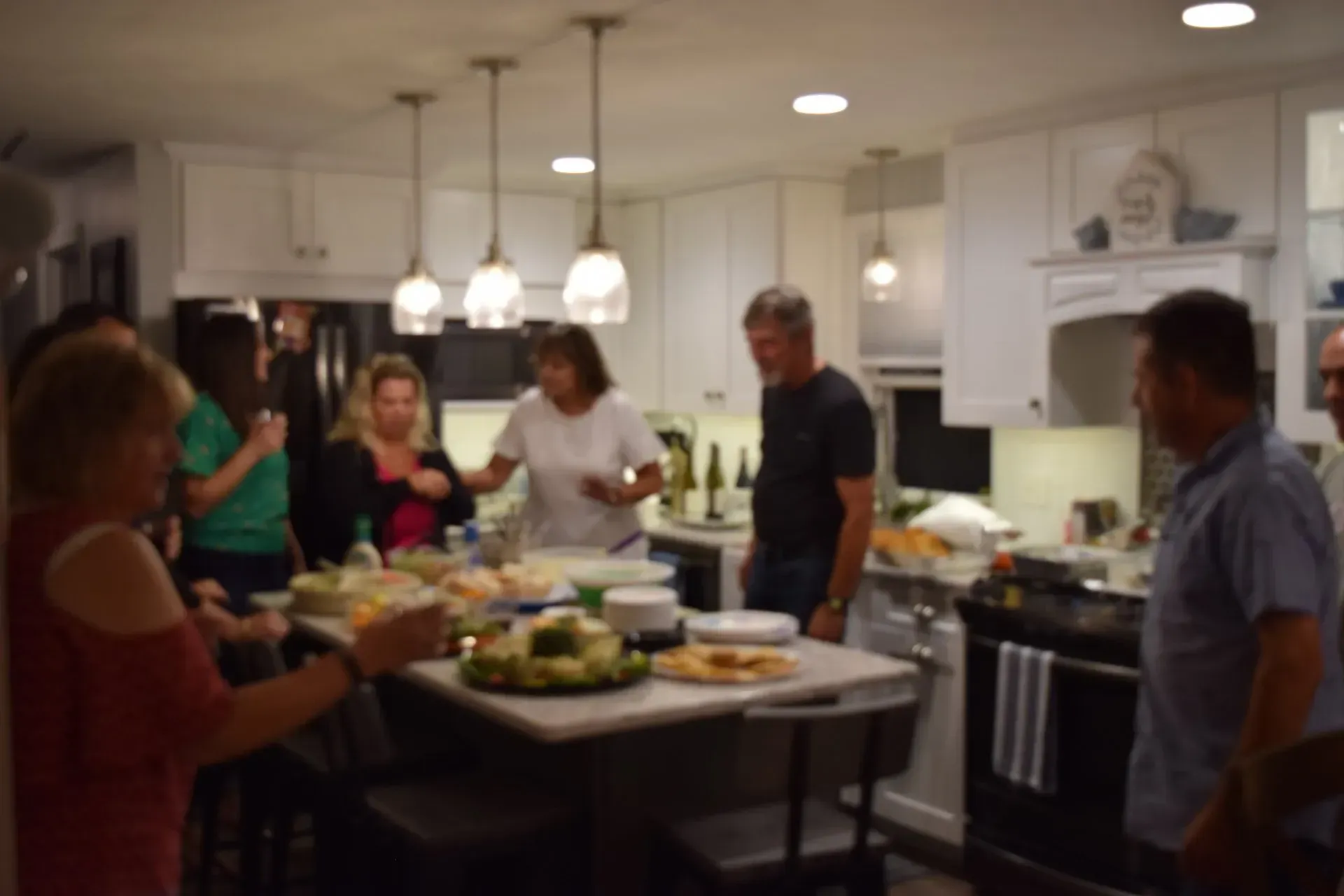 People gathered in a well-lit kitchen, preparing and eating food at an island counter, white cabinets.