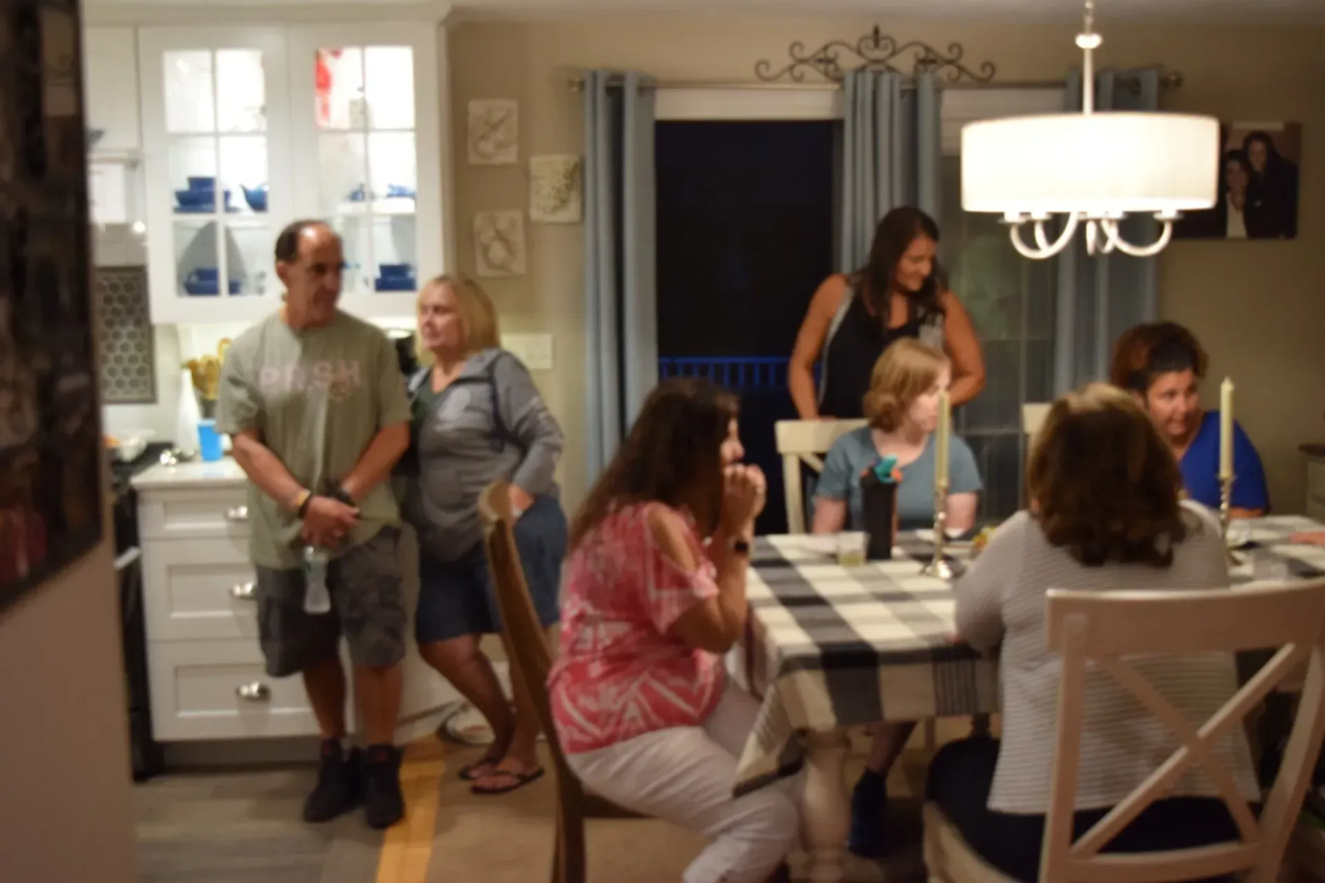 People gather around a table, a kitchen, and doorway. Table setting with a checkerboard tablecloth. Lit room, dark outside.